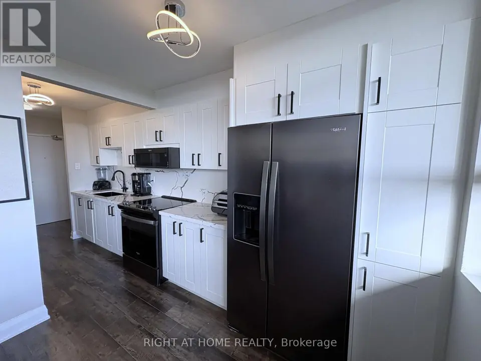 Long kitchen with dark side-by-side refrigerator, tall white cabinets, range and microwave, dark laminate floor leading to a hallway door.