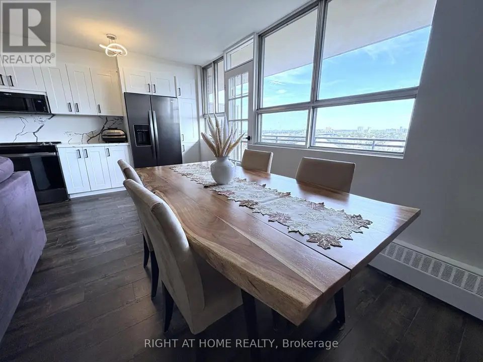 Live-edge dining table in foreground, white kitchen cabinets and dark double-door fridge behind, large windows with city view at right.