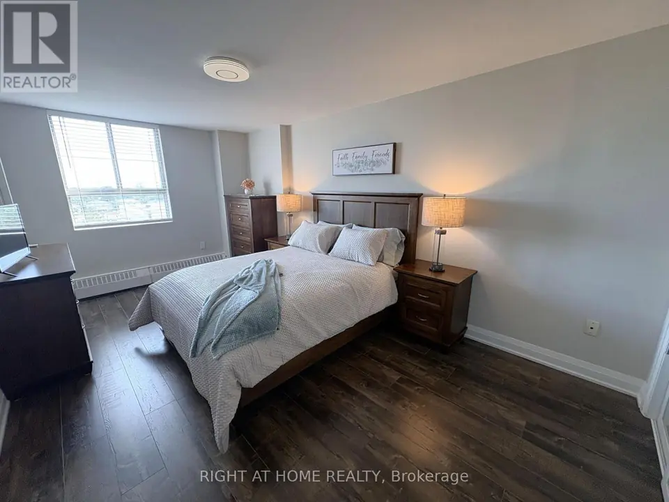 Bedroom with wooden bed, two lit bedside lamps, tall dresser, window with blinds and dark laminate flooring.